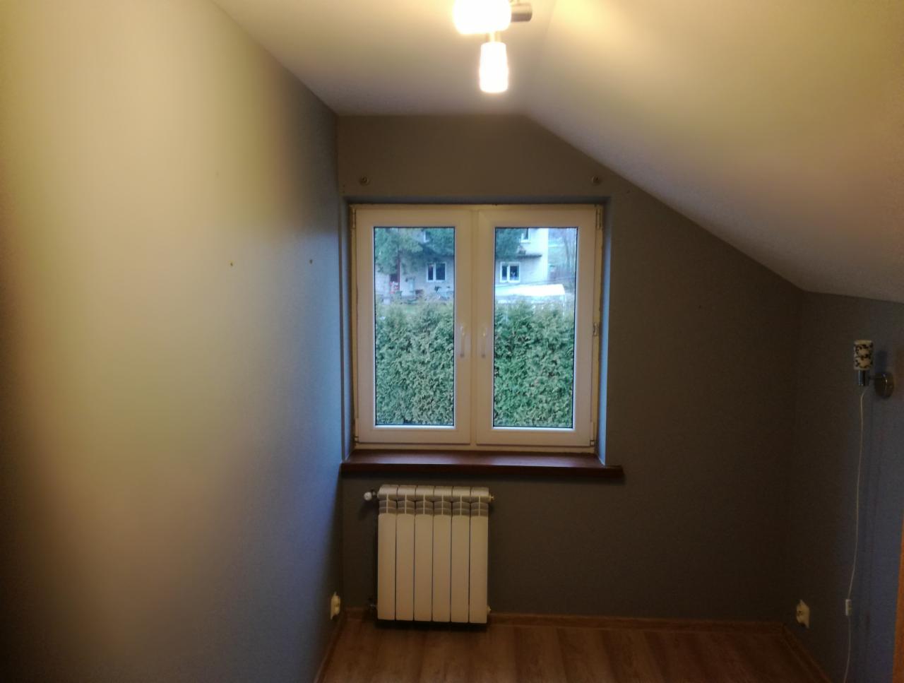 Pokój na poddaszu po remoncie: szare ściany, drewniana podłoga, białe okno z widokiem na zieleń, biały grzejnik pod oknem, kinkiet na ścianie.