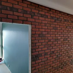 Ściana z cegły klinkierowej w odcieniach czerwieni i brązu, z widocznym przejściem do pomalowanej na niebiesko ściany w sąsiednim pomieszczeniu, jasna podłoga i okno w tle.