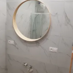 Nowoczesna łazienka z okrągłym lustrem w drewnianej ramie, marmurowe płytki na ścianach, widoczne przyłącza wodne i instalacja prysznicowa w folii ochronnej.