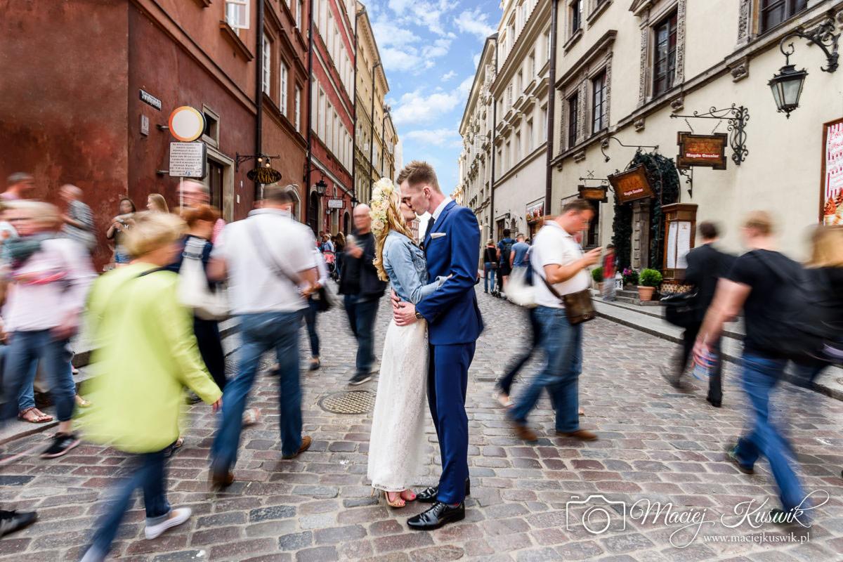 Para młoda całuje się na brukowanej ulicy Starego Miasta w Warszawie, otoczona rozmytymi postaciami przechodniów.