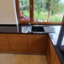 Czarny, błyszczący blat kuchenny z granitu Galaxy Star, zamontowany w kuchni pod oknem z widokiem na zieleń. Widoczny zlew i bateria w kolorze czarnym.