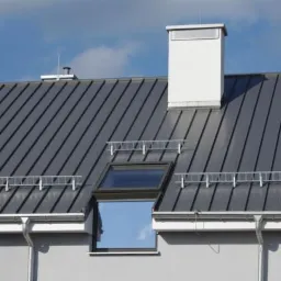Szary dach z blachy na rąbek stojący z oknem dachowym, kominem i systemem odśnieżania, w tle błękitne niebo.