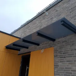 Szklany daszek nad wejściem do budynku z ceglaną elewacją, wsparty na czarnych, metalowych wspornikach. Widoczne przewody elektryczne zwisające obok drzwi.