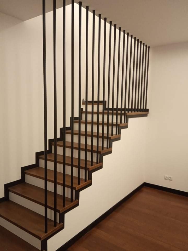 Nowoczesne schody z drewnianymi stopniami i minimalistyczną, czarną, pionową konstrukcją zabezpieczającą zamiast tradycyjnej balustrady, na tle białej ściany.