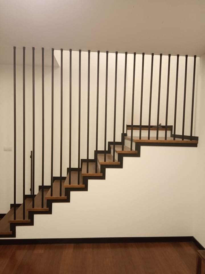 Schody z drewnianymi stopniami i minimalistyczną, pionową barierką z czarnych prętów, widok z dołu na jasnym tle ściany.