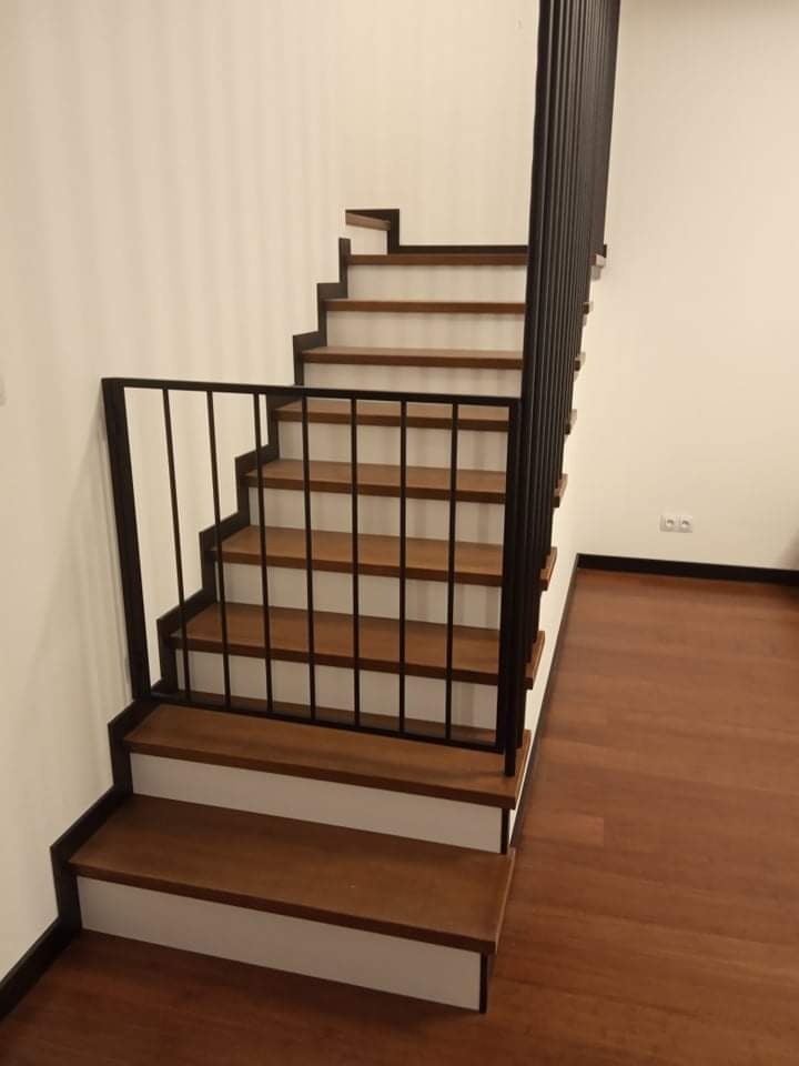 Schody drewniane z czarną, metalową barierką zabezpieczającą na dolnym biegu, widok z dołu na jasne ściany i drewnianą podłogę.