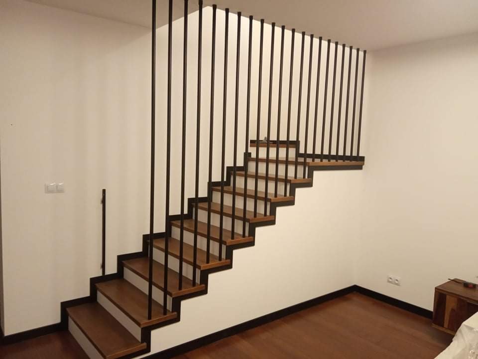 Nowoczesne schody z drewnianymi stopniami i minimalistyczną, czarną balustradą pionową, widoczne z boku na tle białej ściany.