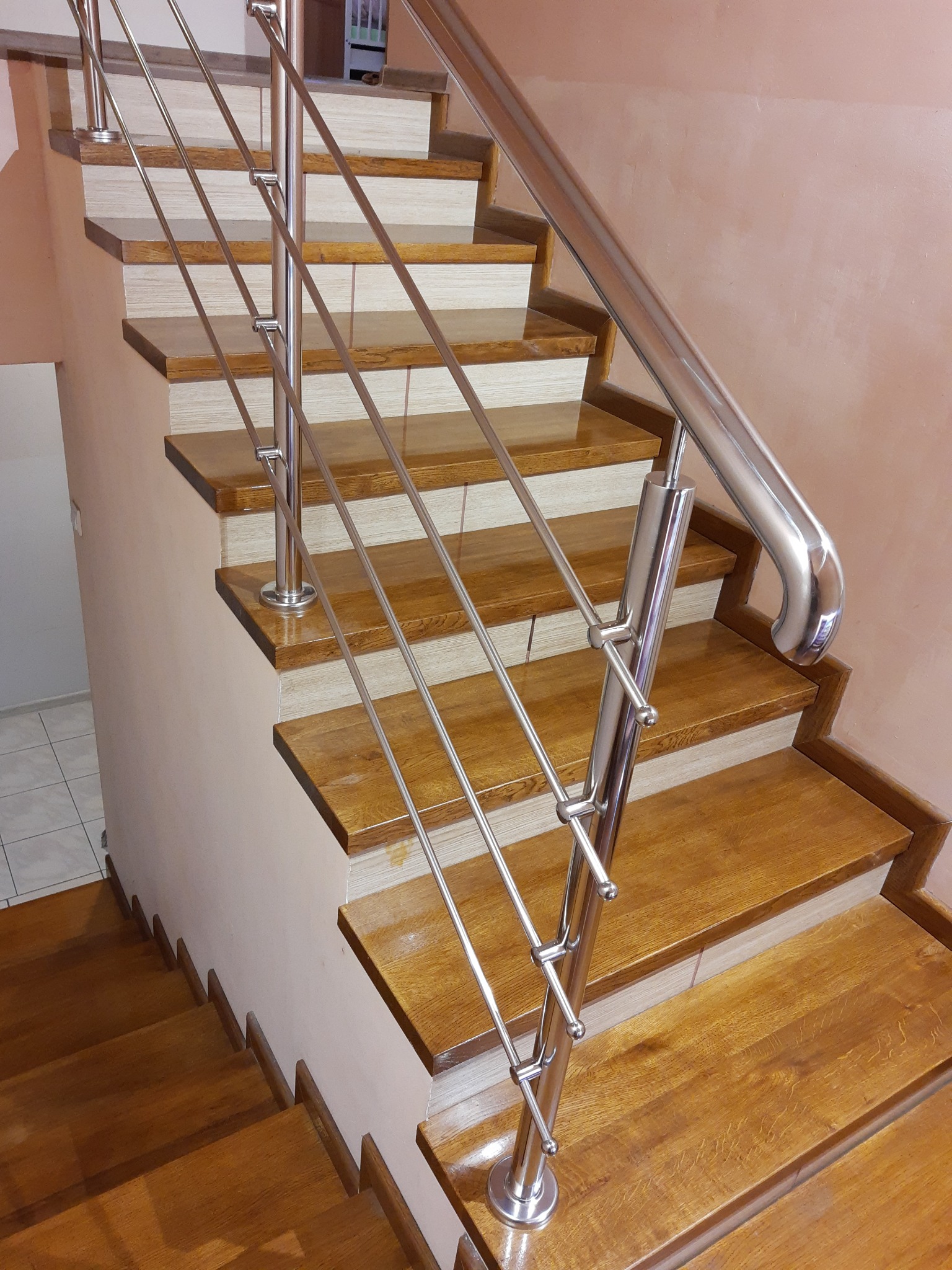 Schody drewniane z błyszczącą metalową barierką, zabezpieczoną poziomymi prętami, widoczne z dołu w kierunku góry.