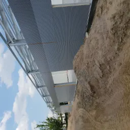 Konstrukcja budynku w trakcie powstawania, widoczny szkielet stalowy i szare płyty z profilowanego metalu, obok hałda ziemi.