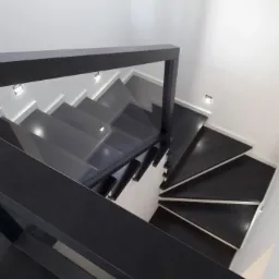 Nowoczesne schody spiralne z czarnymi stopniami i minimalistyczną, czarną balustradą, oświetlone punktowymi lampami w ścianie, widok z góry.