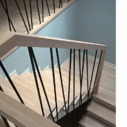 Nowoczesne schody wewnętrzne z jasnego drewna z czarnymi, pionowymi balustradami, widok z góry na dół, ściana w tle w odcieniu szaro-niebieskim.