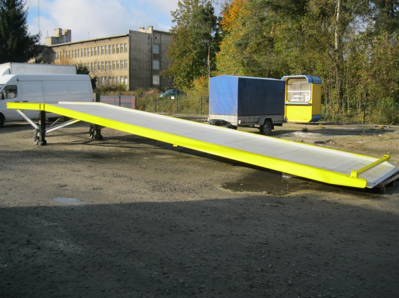 Żółta rampa załadunkowa z perforowaną powierzchnią, ustawiona na zewnątrz, w tle budynek i drzewa.