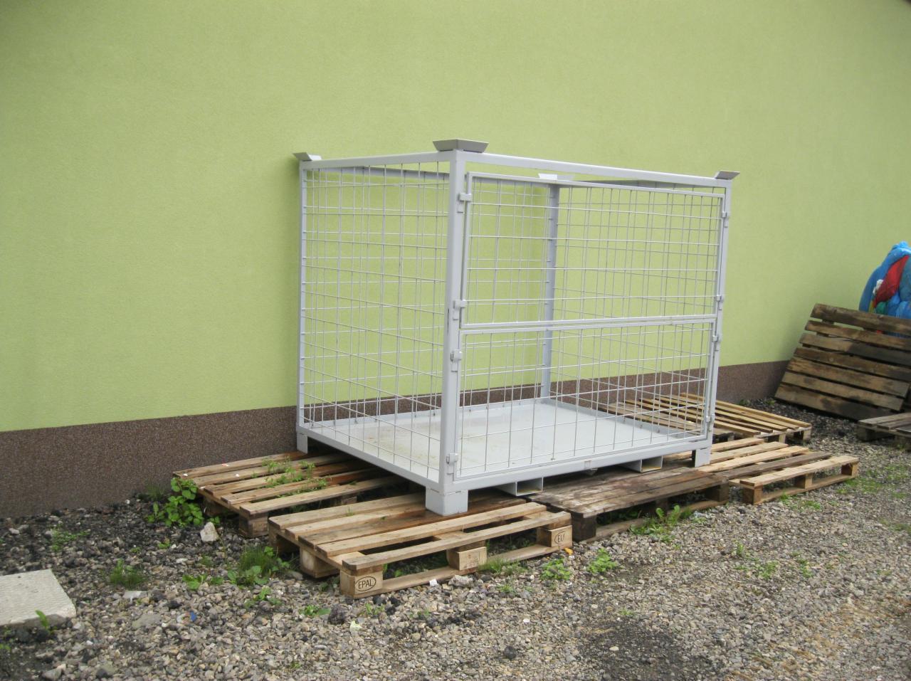 Klatka transportowa metalowa, siatkowa, postawiona na paletach przed zieloną ścianą budynku.