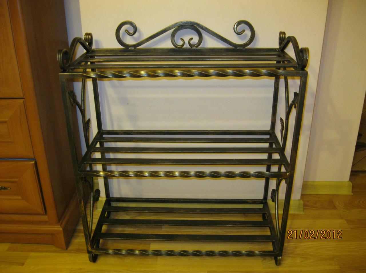 Trzypoziomowy, metalowy regał na buty z kutymi zdobieniami w kształcie spiral i liści, stojący na drewnianej podłodze przy ścianie.