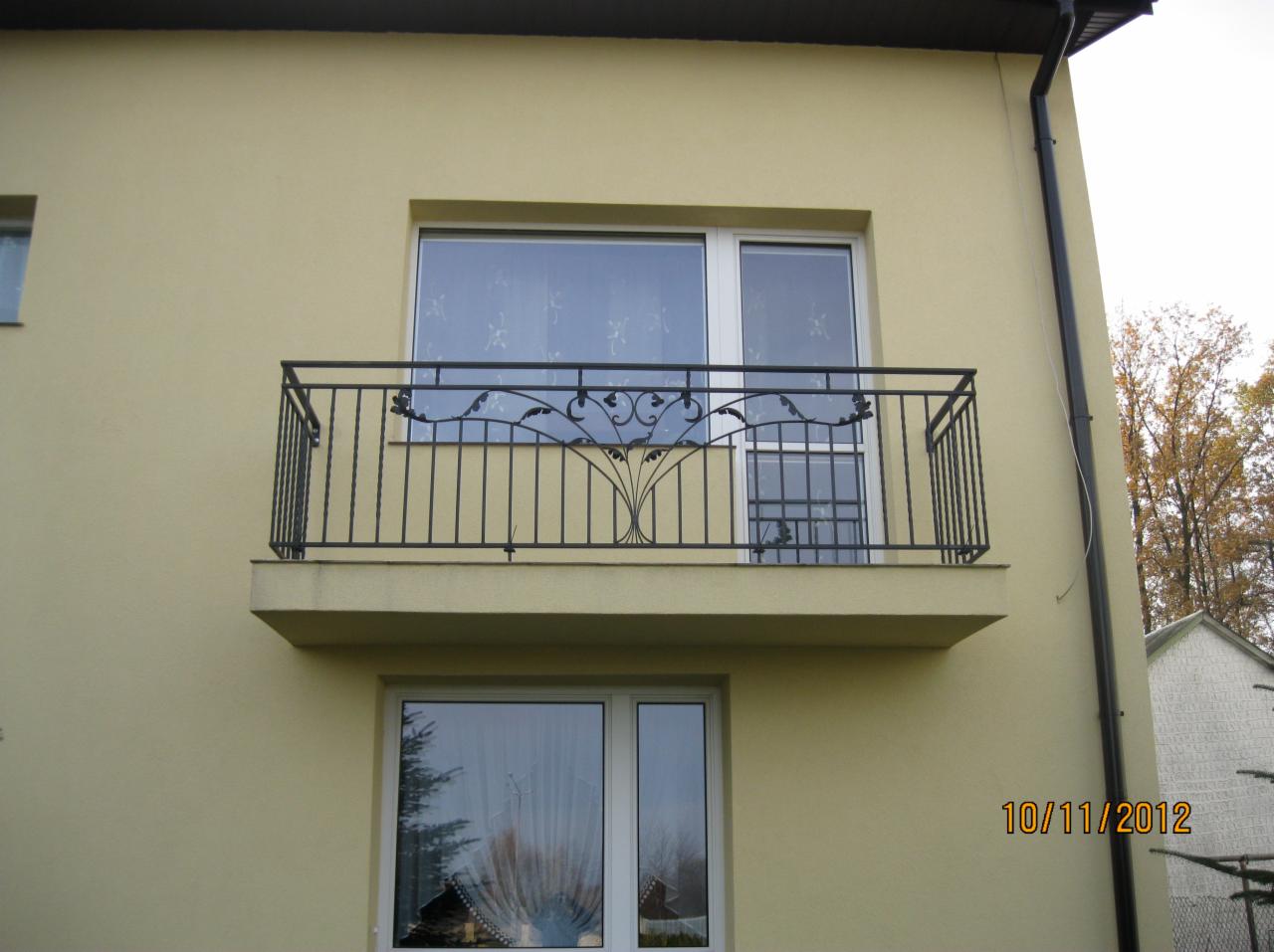 Balkon z czarną metalową balustradą o dekoracyjnym, stylizowanym na roślinny wzorze, zamontowaną na beżowej elewacji budynku.