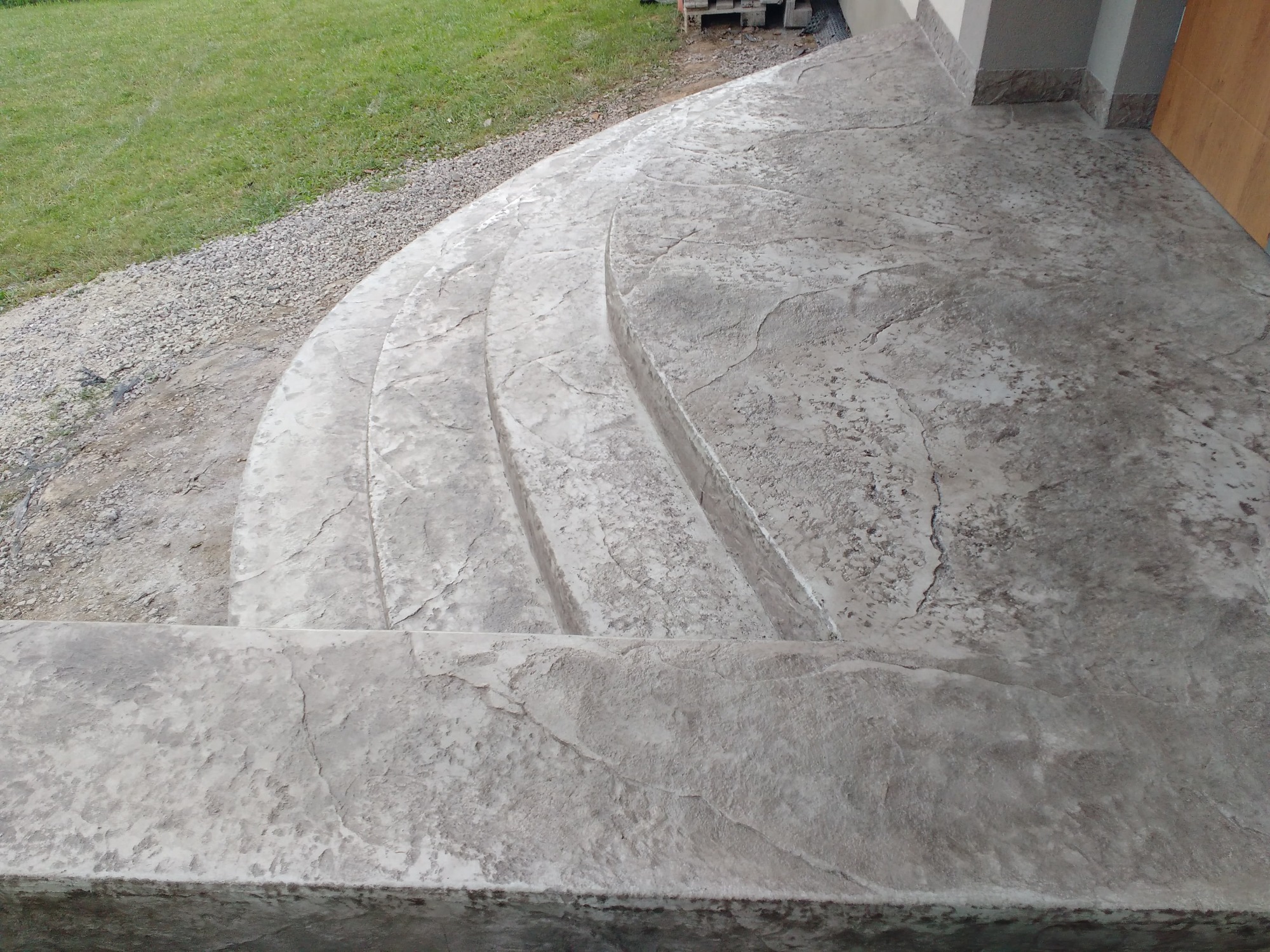 Wykończone schody zewnętrzne z betonu stemplowanego, imitującego kamień, prowadzące do drzwi wejściowych, z widocznym otoczeniem trawnika i podsypki żwirowej.