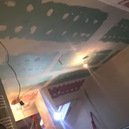 Sufit w trakcie szpachlowania z widocznymi łatami i prowizorycznym oświetleniem żarówkowym, ukazujący etap prac wykończeniowych przed malowaniem.