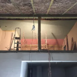 Poddasze w trakcie prac wykończeniowych: widoczna izolacja wełną mineralną pod dachem, szare płyty gipsowo-kartonowe na suficie, drabina i rusztowanie w pomieszczeniu, surowe ściany w odcieniach...