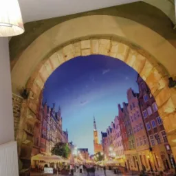 Fototapeta na ścianie przedstawiająca ulicę w Gdańsku widzianą przez kamienny łuk, widoczny grzejnik i fragment podłogi.
