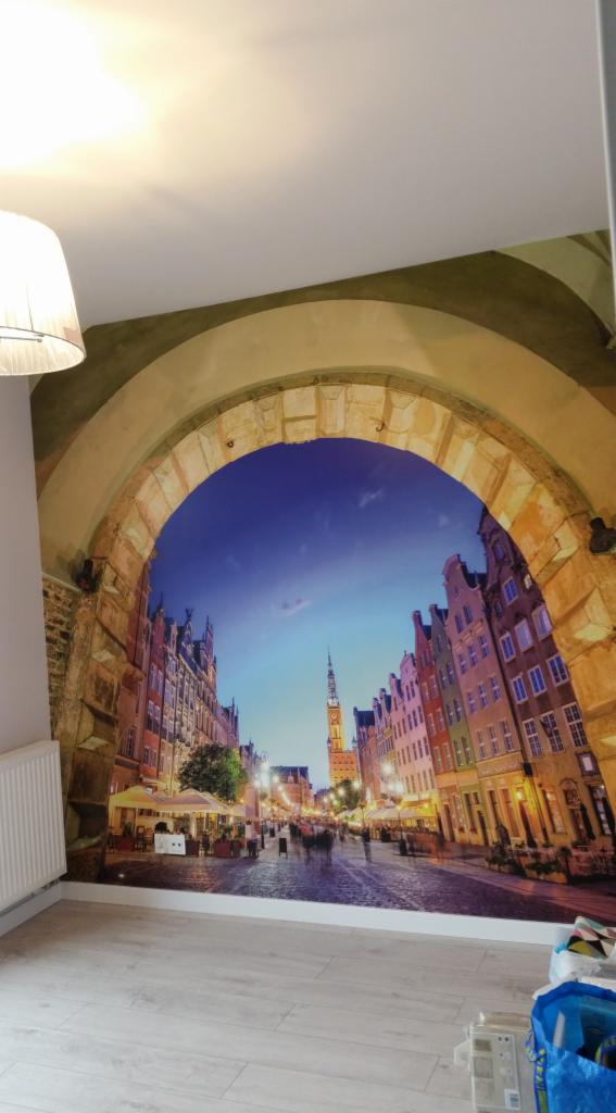 Fototapeta na ścianie przedstawiająca ulicę w Gdańsku widzianą przez kamienny łuk, widoczny grzejnik i fragment podłogi.