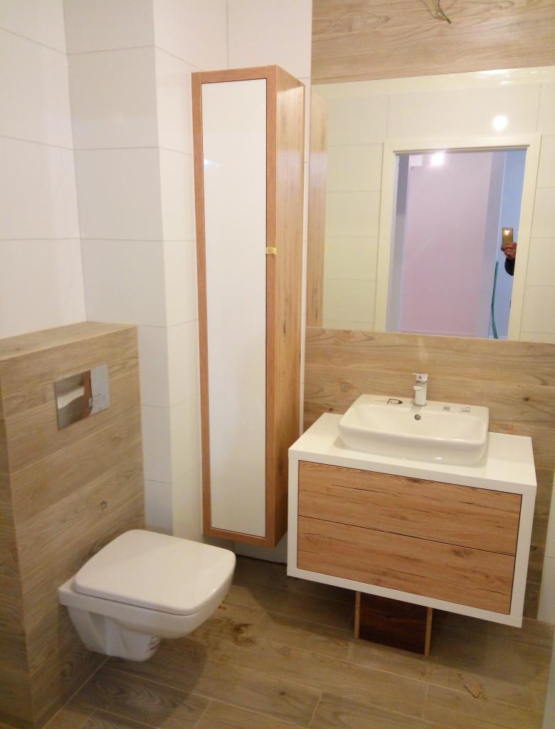 Nowoczesna łazienka z podwieszaną toaletą, szafką z umywalką z drewnianymi frontami i wysoką szafką, wykończona płytkami imitującymi drewno.