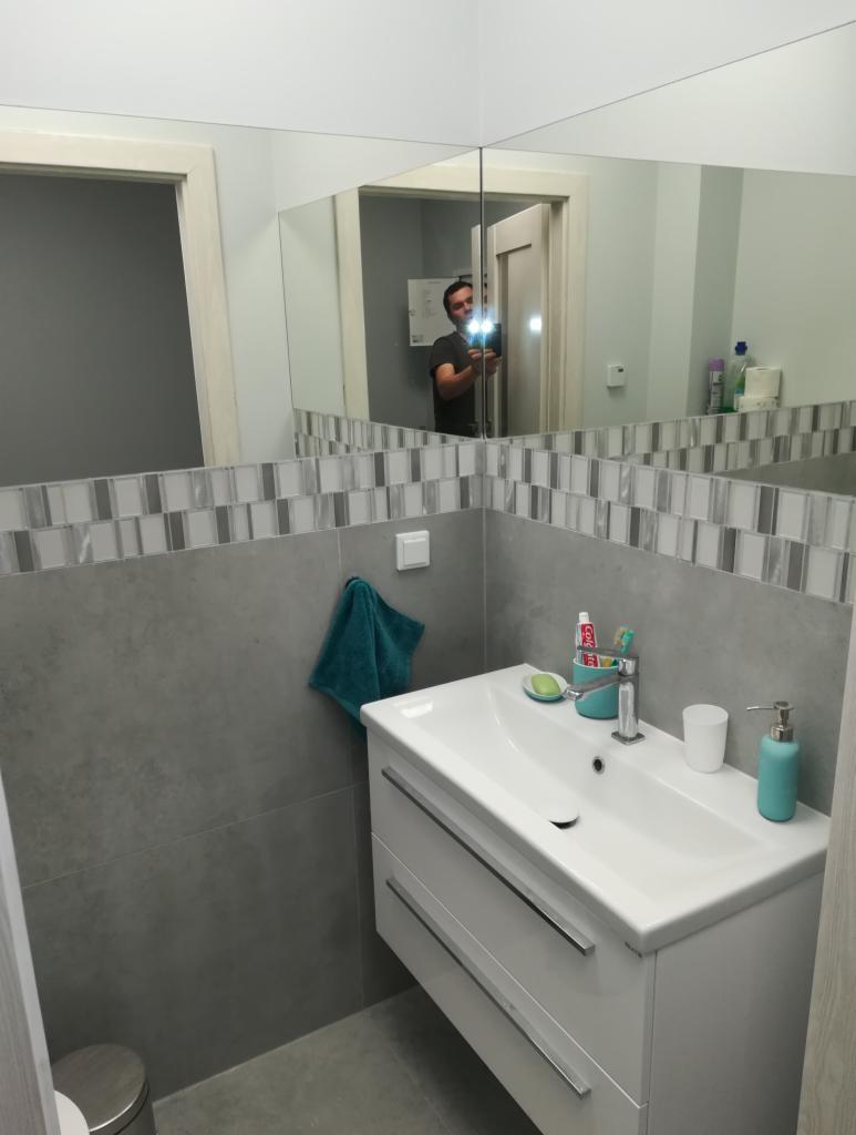 Szara łazienka po remoncie z umywalką nablatową, białą szafką z dwoma szufladami, mozaiką na ścianie i lustrem, w którym odbija się osoba robiąca zdjęcie.