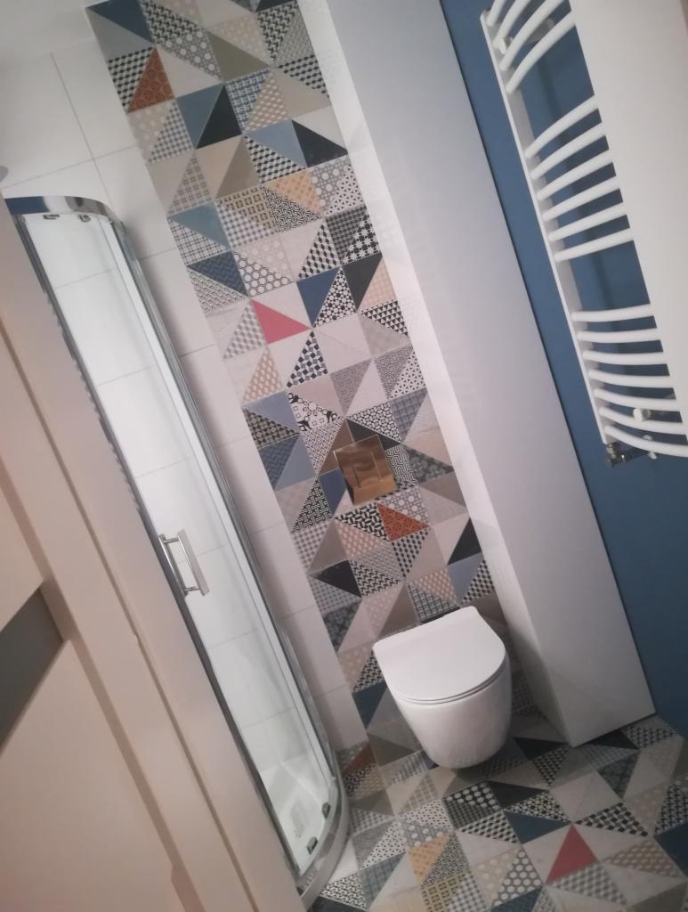 Wykończona łazienka w Komornikach z kabiną prysznicową, toaletą i grzejnikiem drabinkowym. Uwagę przyciągają geometryczne płytki w odcieniach szarości, błękitu i brązu, zdobiące ścianę i podłogę.