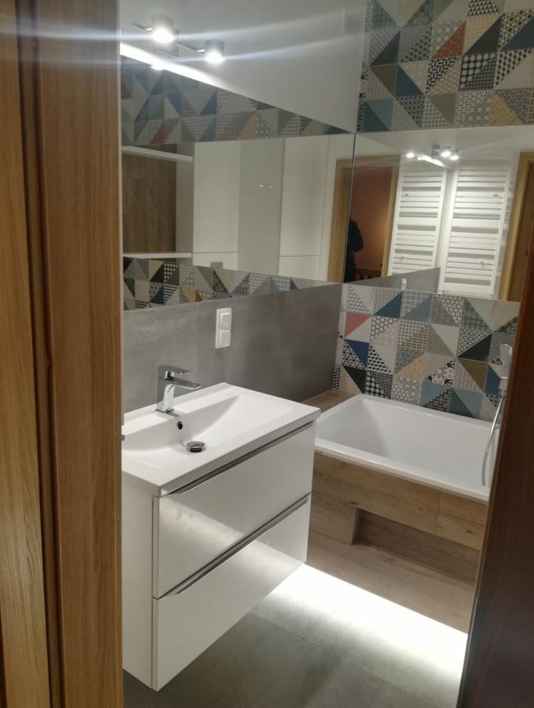 Nowoczesna łazienka z białą szafką pod umywalkę, lustrem odbijającym wzorzyste płytki i wanną z drewnianą obudową, oświetlenie LED pod szafką.