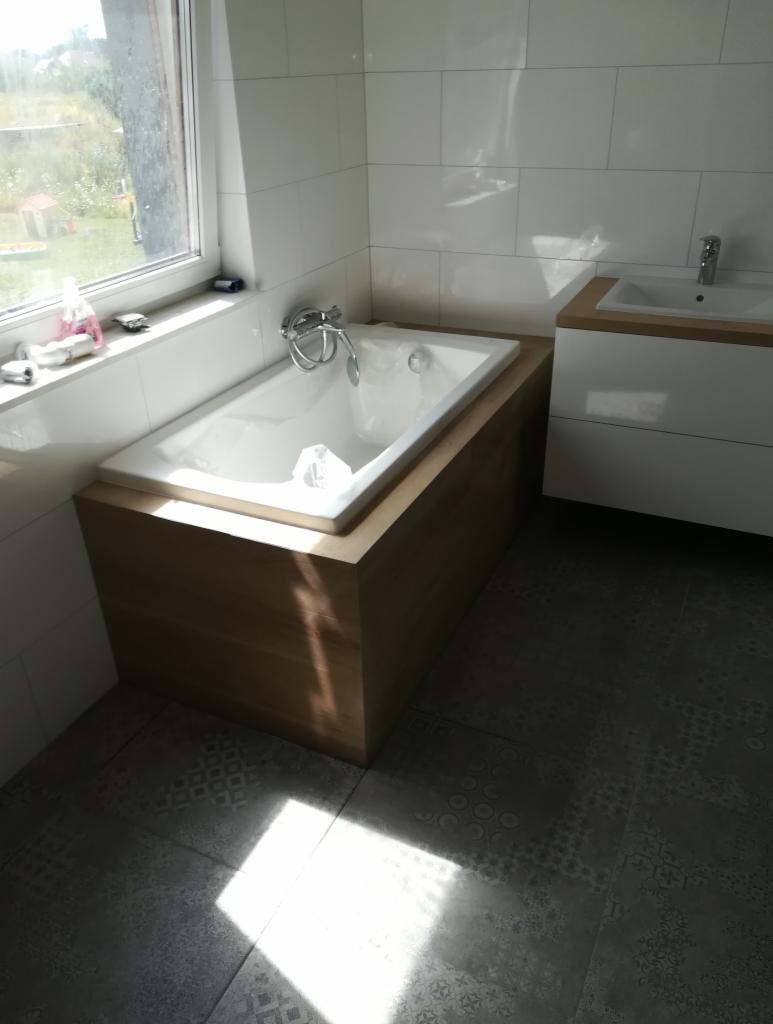 Biała wanna obudowana drewnopodobną płytą, bateria wannowa chromowana, białe płytki na ścianach i szara wzorzysta podłoga w łazience z oknem.