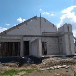 Surowy stan budynku z białych bloczków, widoczne drewniane elementy konstrukcyjne dachu, rusztowanie przy ścianie, teren wokół budynku w trakcie przygotowywania.