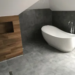 Nowoczesna łazienka na poddaszu z wolnostojącą białą wanną o organicznym kształcie, szarą płytką imitującą beton na podłodze i ścianach oraz akcentem drewnopodobnej okładziny z wnęką.