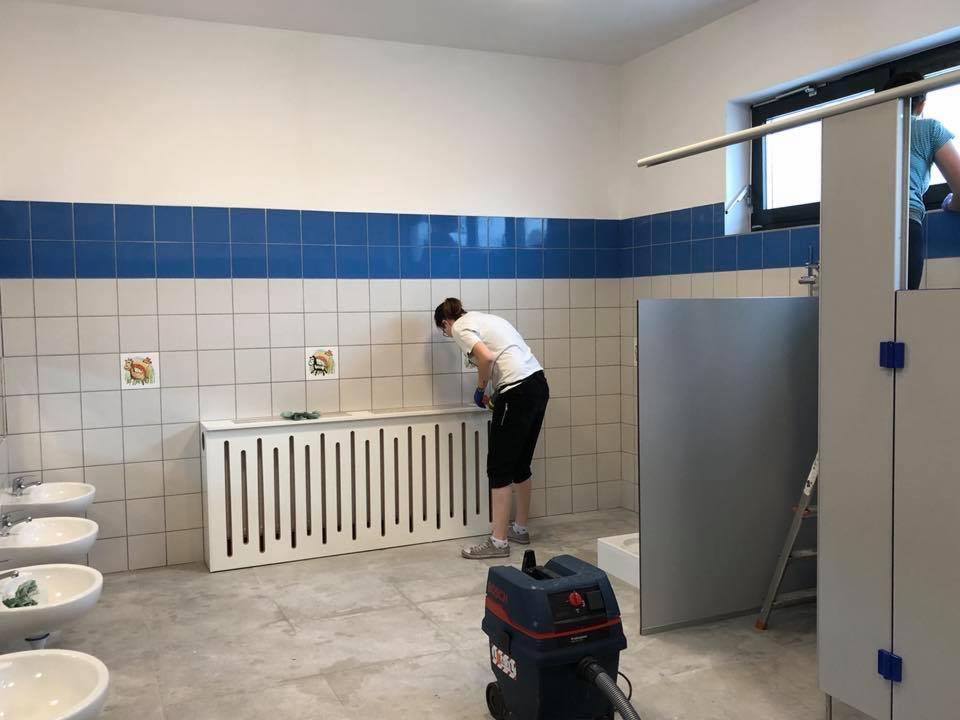 Pracownik w białej koszulce i czarnych spodenkach czyści płytki na ścianie w toalecie publicznej, widoczny odkurzacz przemysłowy Bosch i umywalki.