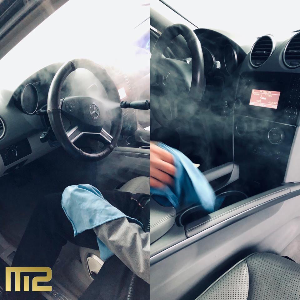 Czyszczenie parowe wnętrza samochodu Mercedes-Benz, widoczne para i niebieska ściereczka z mikrofibry, logo firmy w lewym dolnym rogu.