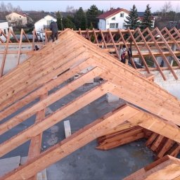 RoofExpert Dachy - Drewniana konstrukcja dachu w trakcie montażu, widoczne elementy więźby dachowej, dwóch pracowników na dachu, domy w tle.