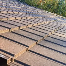 Nowe, brązowe pokrycie dachowe z blachy trapezowej, widoczne deski więźby dachowej i fragment krajobrazu z drzewami iglastymi w tle.