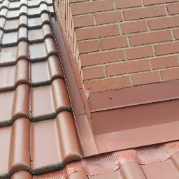 Czerwona dachówka ceramiczna z obróbką blacharską komina z cegły, widoczne zabrudzenia na dachówkach.