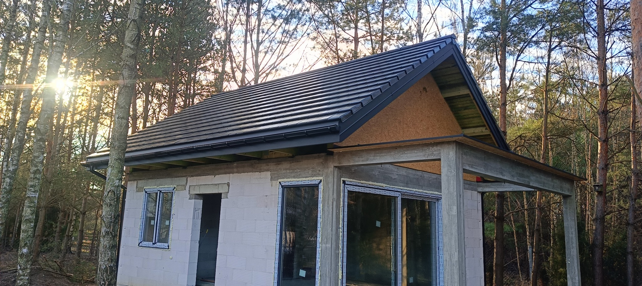 Surowy stan budynku z czarną dachówką na tle leśnym, widoczne okna z pianką montażową i niezabezpieczona elewacja z betonu komórkowego.