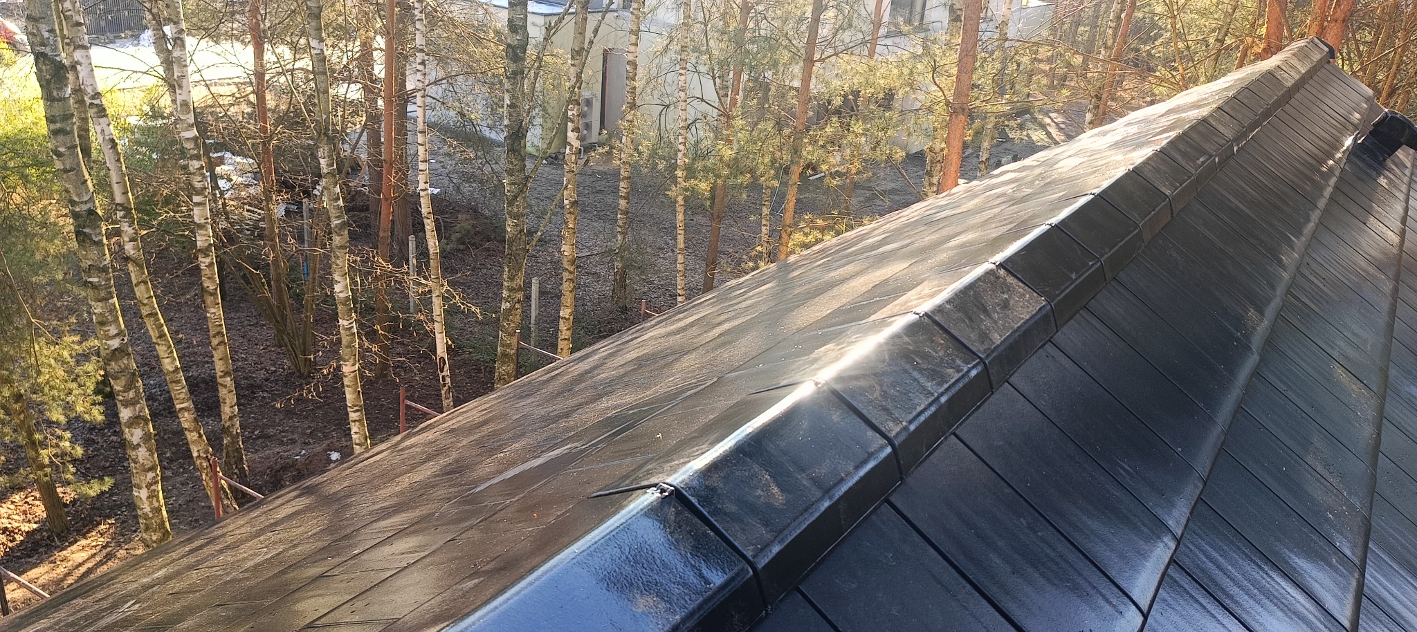 Nowy, błyszczący, czarny dach z dachówki ceramicznej w promieniach słońca, widok z góry na tle drzew i budynku.