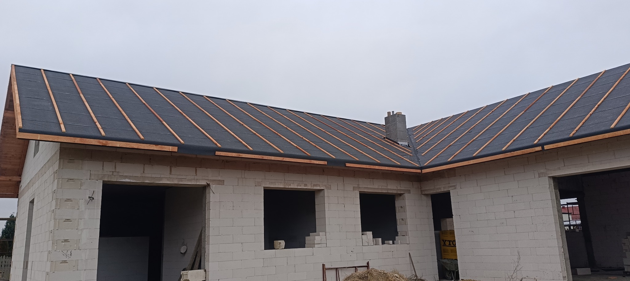 Surowy stan budynku z białego pustaka z widoczną konstrukcją przygotowaną pod pokrycie dachu papą i łatami, widoczny komin