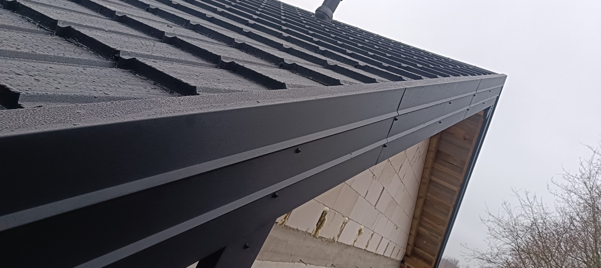 Nowo położone, grafitowe pokrycie dachowe z blachy na skośnym dachu, widoczne krople wody, obróbki blacharskie przy okapie, surowy stan budynku z widoczną konstrukcją drewnianą i fragmentem muru...