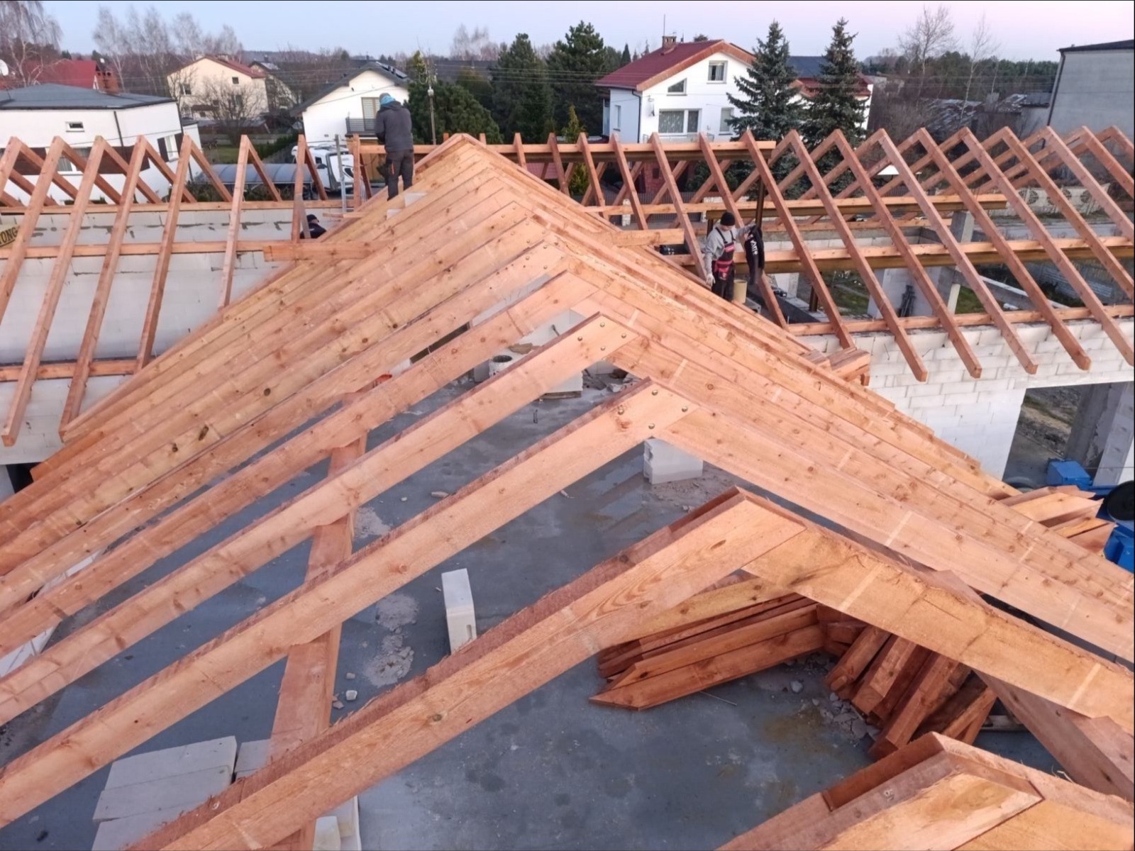 Drewniana konstrukcja dachu w trakcie montażu, widoczne elementy więźby dachowej, dwóch pracowników na dachu, domy w tle.