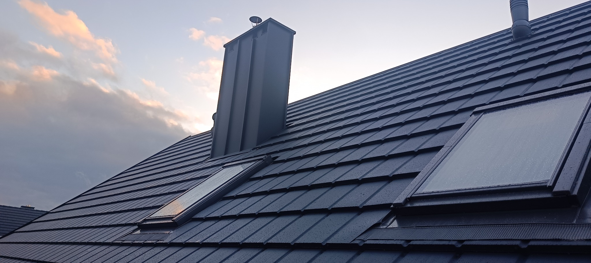 Nowy, grafitowy dach z dachówki ceramicznej z dwoma oknami dachowymi i kominem, widoczne krople deszczu, wieczorne niebo w tle.