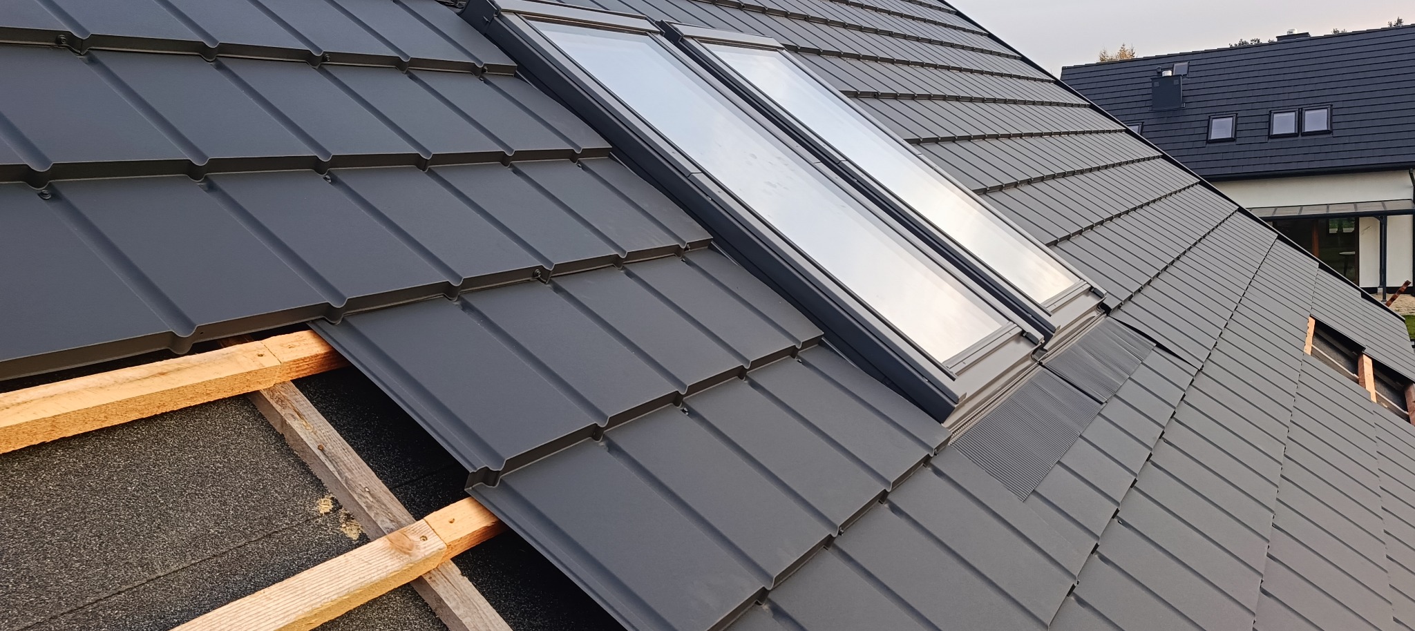 Montaż okien dachowych na nowym, grafitowym pokryciu z blachodachówki, widoczna konstrukcja więźby i podkład z papy.