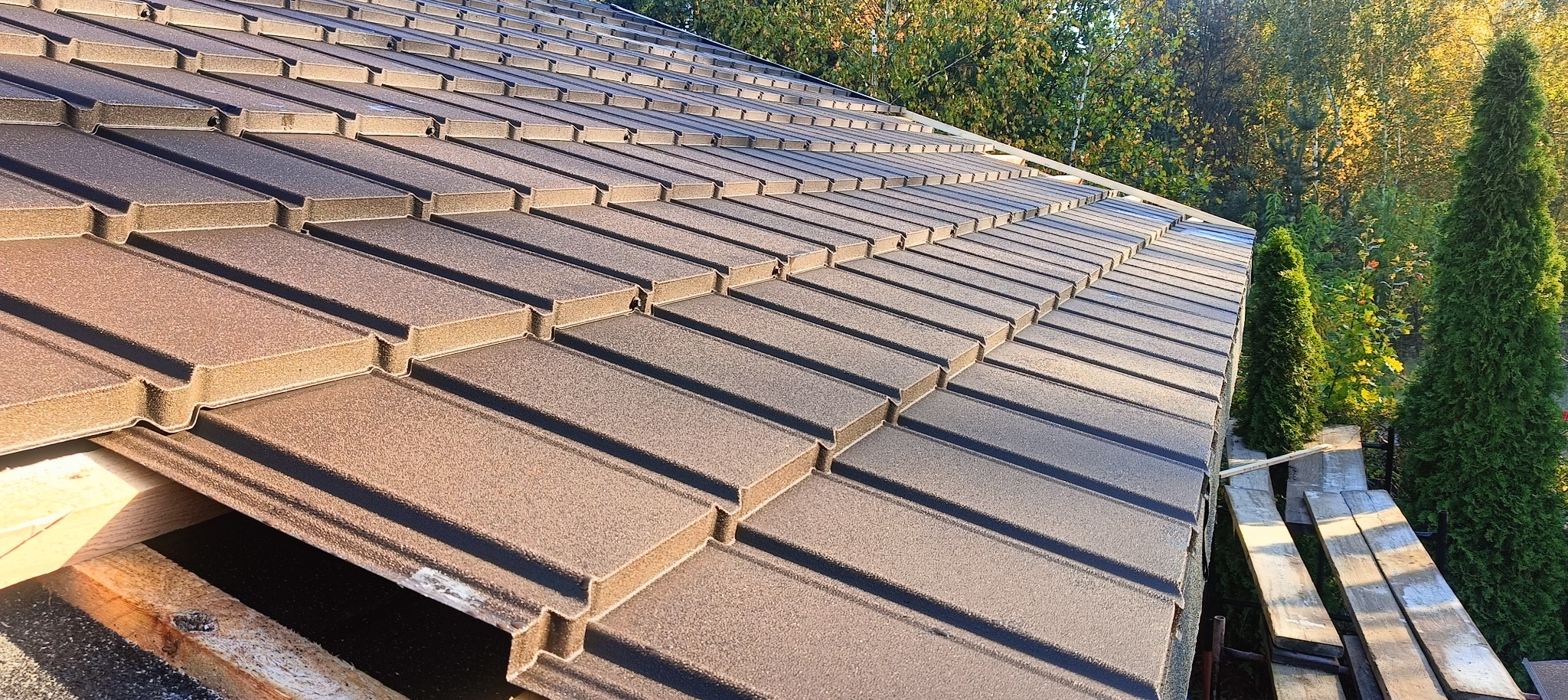 Nowe, brązowe pokrycie dachowe z blachy trapezowej, widoczne deski więźby dachowej i fragment krajobrazu z drzewami iglastymi w tle.