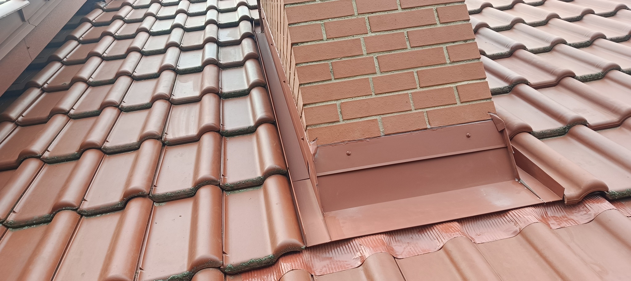 Czerwona dachówka ceramiczna z obróbką blacharską komina z cegły, widoczne zabrudzenia na dachówkach.