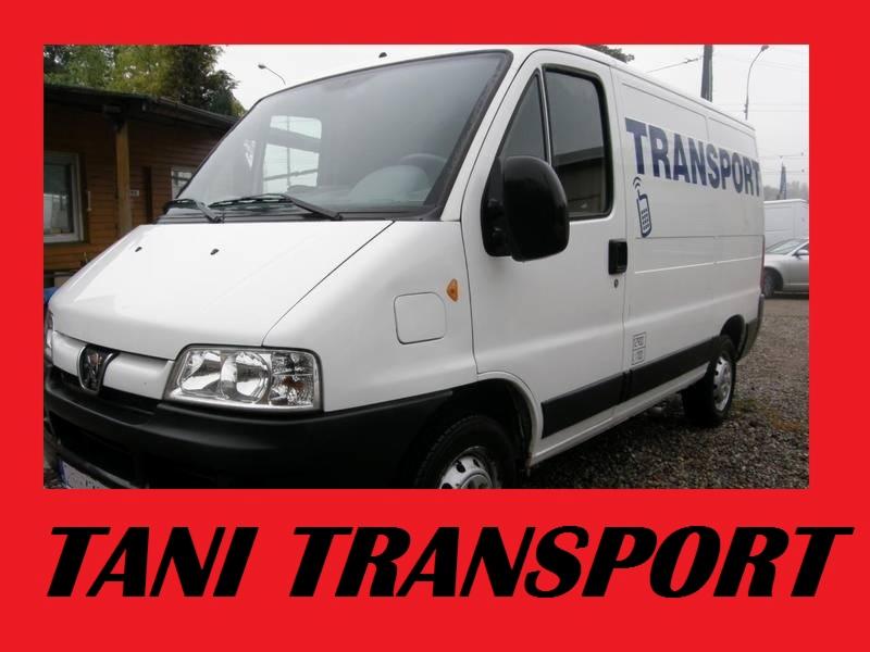 Biały samochód dostawczy z napisem TRANSPORT i symbolem telefonu komórkowego na boku, zaparkowany przed budynkiem.