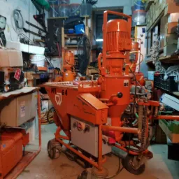 Pomarańczowa, mobilna maszyna PFT G4 do nakładania tynków, widoczna w warsztacie z narzędziami i materiałami budowlanymi.