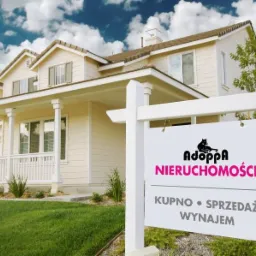 Dom jednorodzinny z beżową elewacją i białymi oknami, na pierwszym planie tablica agencji nieruchomości z logo czarnego kota i napisem 'Kupno &bull; Sprzedaż &bull; Wynajem'.