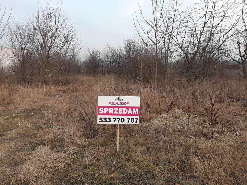 Tablica z napisem 'Sprzedam' i numerem telefonu na tle zarośniętej działki w pochmurny dzień.