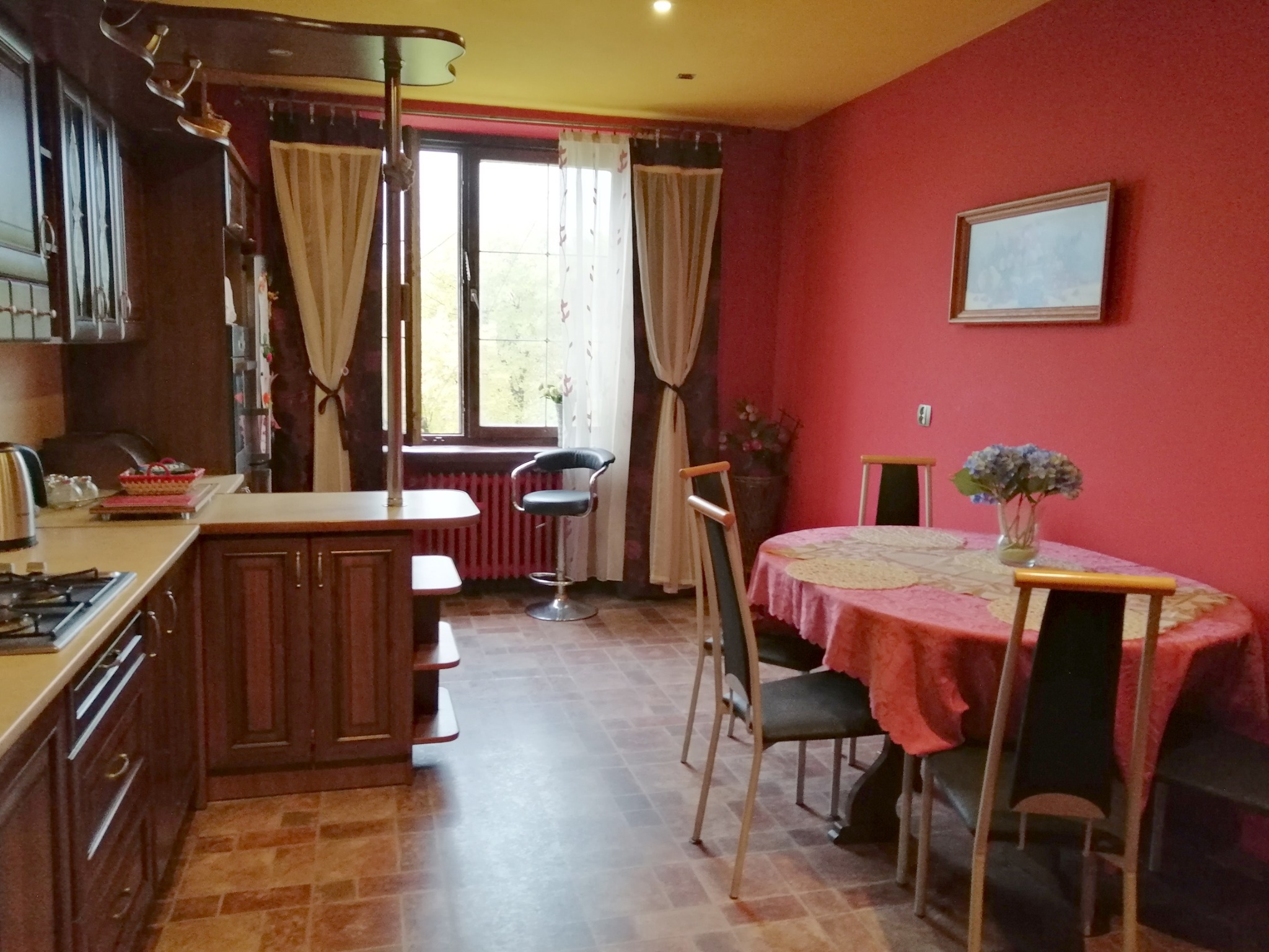 Umeblowana kuchnia w odcieniach czerwieni i brązu z oknem z widokiem na zieleń, stół z kwiatami i krzesłami oraz bar z oświetleniem.